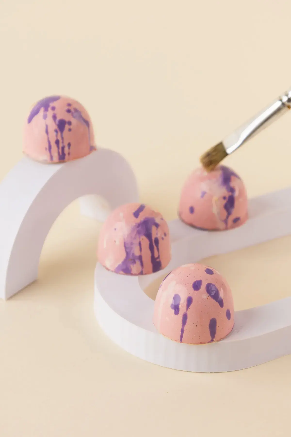 Pincel pintando de roxo bombons cor-de-rosa apoiados em suportes brancos